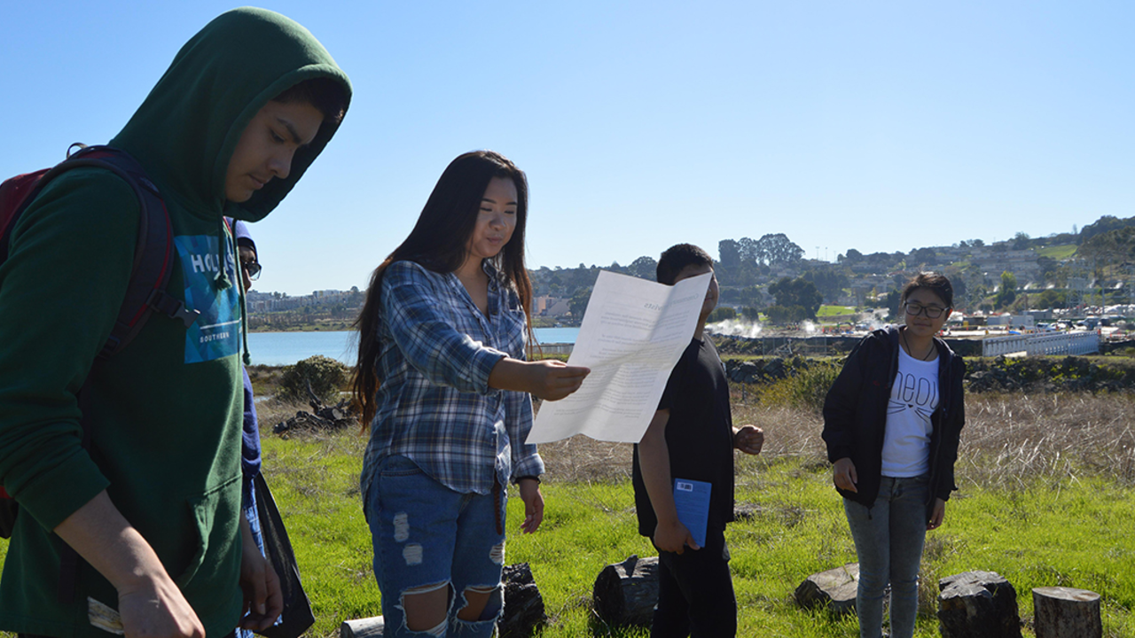 Leading Environmental Science Presentations at Heron's Head A group of four high school students stand in a line outdoors while leading a presentation about the natural environment