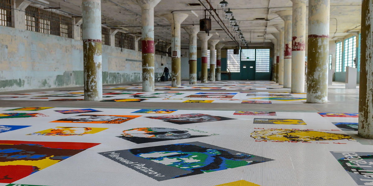Lego brick pieces are arranged to create portraits along a floor inside a large concrete room with columns