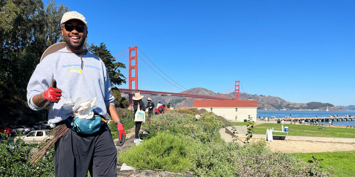 A volunteer gives a thumbs up at Crissy Field in San Francisco with a backdrop of the Golden Gate Bridge and the Warming Hut.