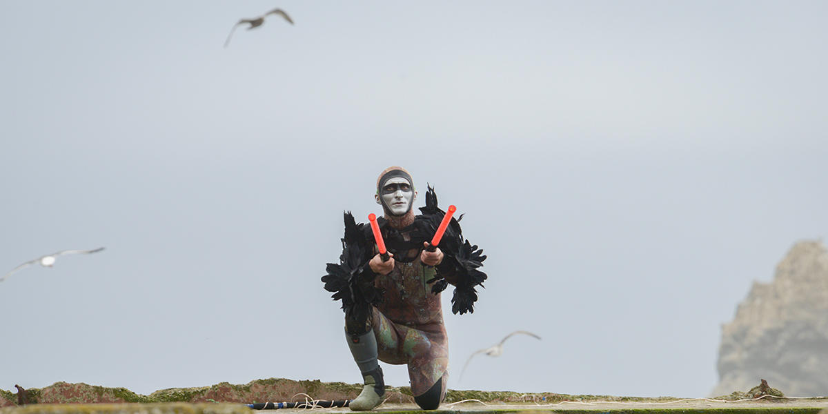 Musician Andy Meyerson wears an elaborate costume on ruins of Sutro Baths with birds flying in background.