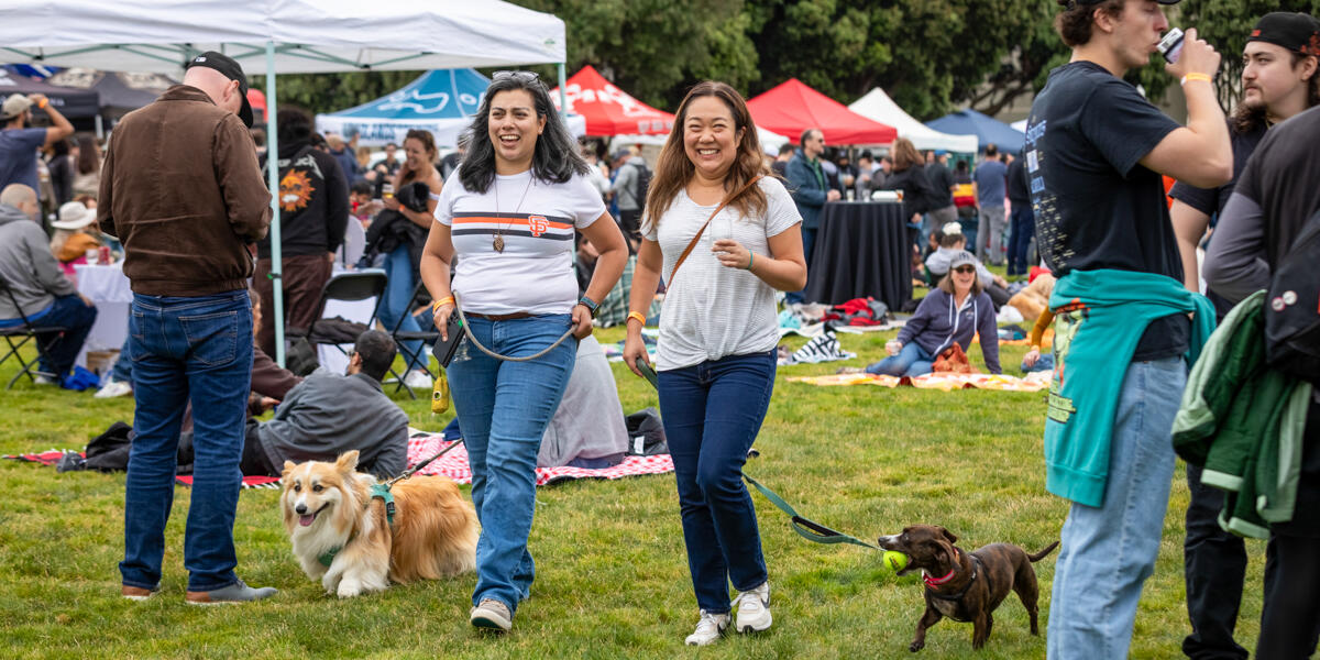 Two women walk their dogs amongst the lawn at Brewfest 2024 in the Presidio