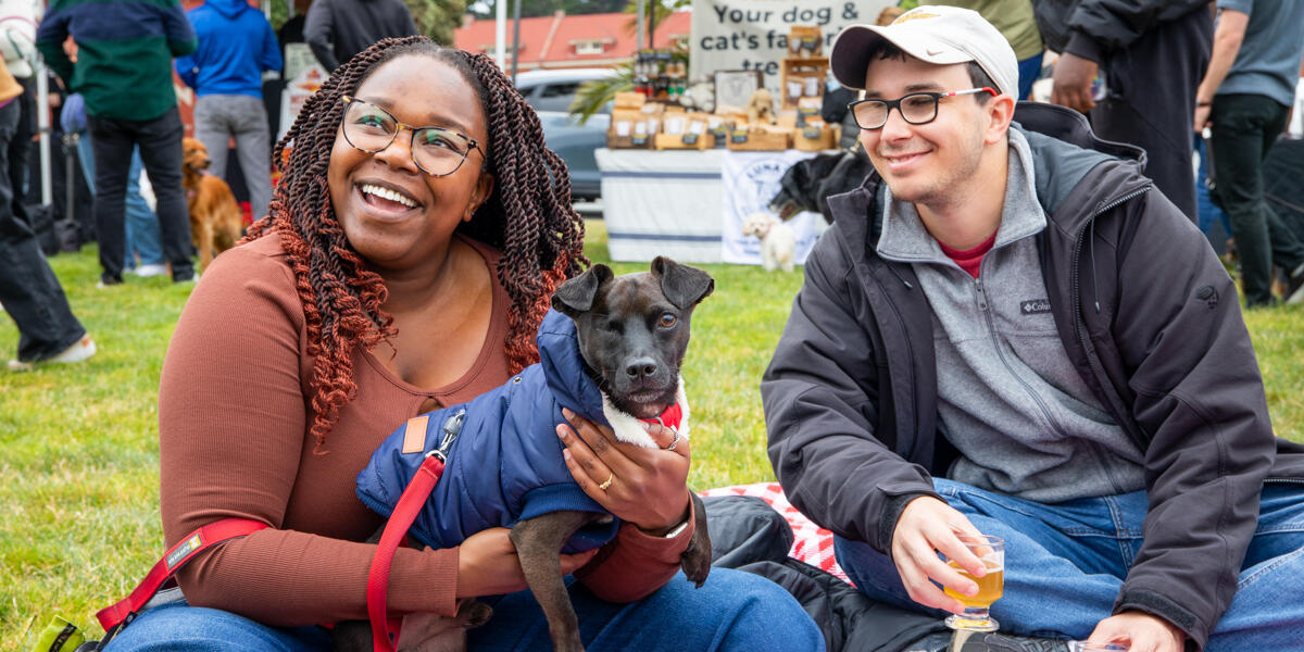 A smiling couple sits with their dog at the Parks4All: Brewfest in the Presidio