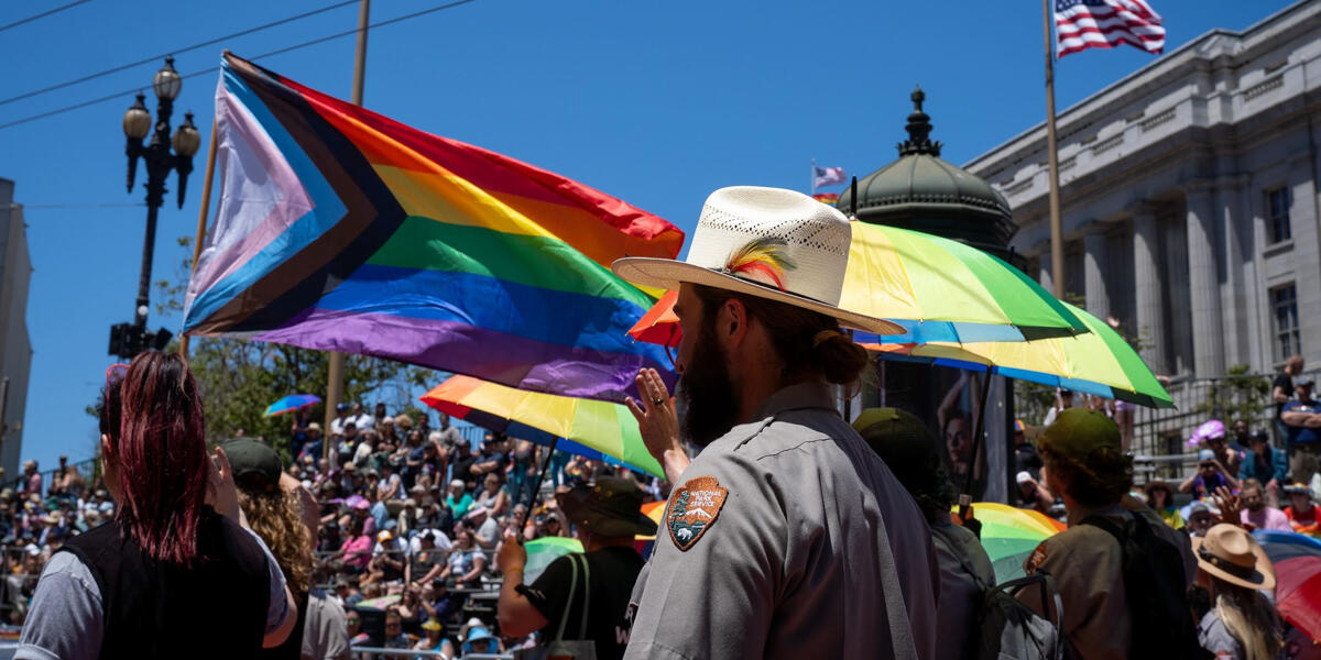 The side of a ranger's hat with a rainbow feather as he marches in the Pride Parade. Rainbow flags and umbrellas billow in the background.