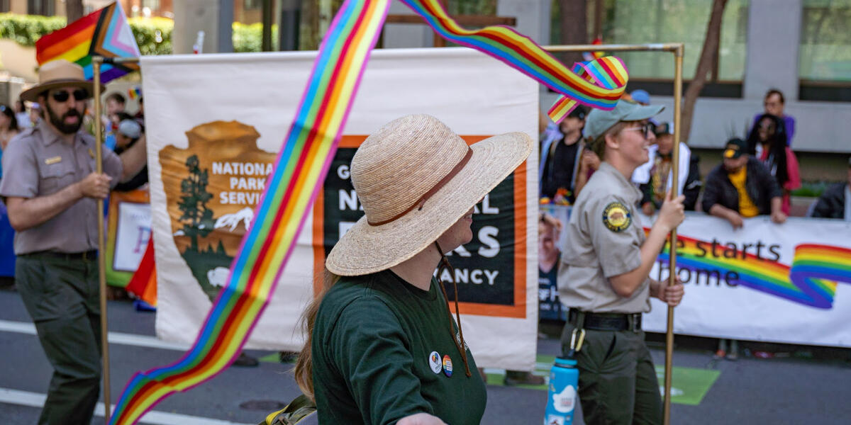 A ranger in a straw hat swirls a rainbow ribbin, while two other rangers hold a Golden Gate National Parks banner in the background.