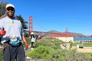 A volunteer gives a thumbs up at Crissy Field in San Francisco with a backdrop of the Golden Gate Bridge and the Warming Hut.