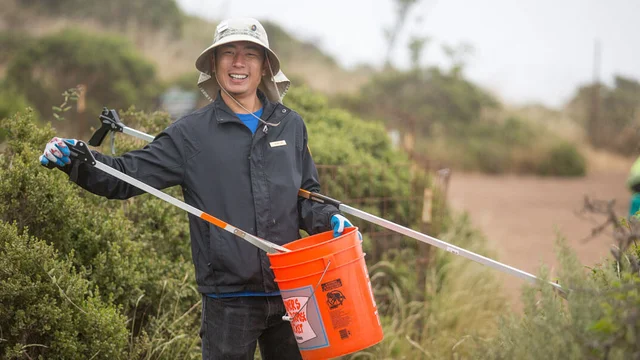 A volunteer holds a bucket and debris grabbing tool on a trail.