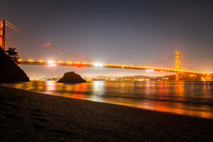 A night view of the Golden Gate Bridge from Kirby Cove. The lights from the bridge glow on the water and there are stars twinkling overhead.