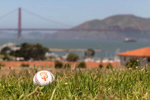A baseball with the SF Giants logo lays on the green lawn at Fort Mason with a view of the Golden Gate Bridge in the background.
