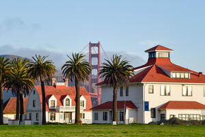 A view from Crissy Field looking out through buildings and palm trees at the Golden Gate Bridge.