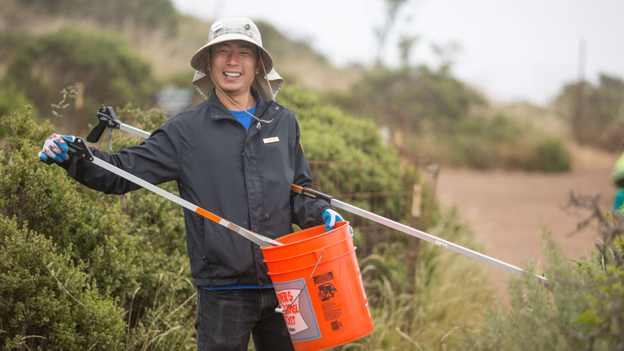 A volunteer holds a bucket and debris grabbing tool on a trail. A volunteer holds a bucket and debris grabbing tool on a trail.