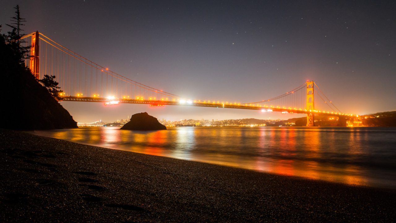 A night view of the Golden Gate Bridge from Kirby Cove. A night view of the Golden Gate Bridge from Kirby Cove. The lights from the bridge glow on the water and there are stars twinkling overhead.
