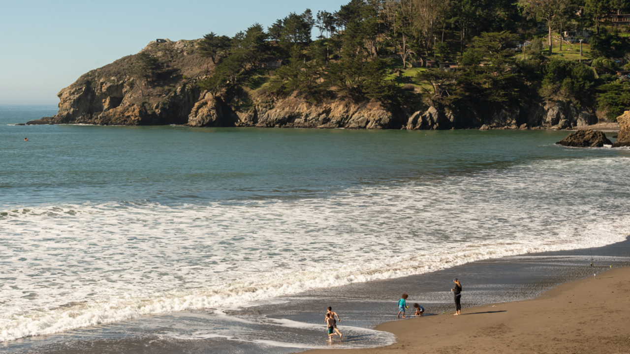 A Picturesque Scene at Muir Beach Children play at the waters edge while a parent watches at Muir Beach. Tree covered mountains are seen jutting out into the ocean just off in the distance.