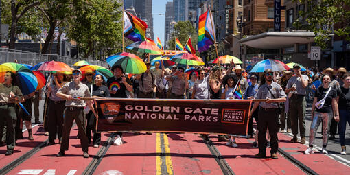Smiling Parks Conservancy staff and NPS Rangers hold a Golden Gate National Parks banner and rainbow umbrellas in the Pride Parade.