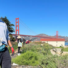 A volunteer gives a thumbs up at Crissy Field in San Francisco with a backdrop of the Golden Gate Bridge and the Warming Hut.